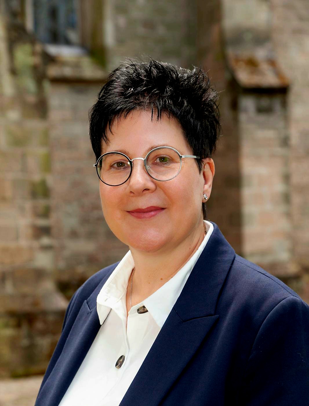 Porträt von Stefanie Kallenborn. Sie trägt eine Brille, hat kurze dunkle Haare und ist in einem dunklen Blazer mit weißer Bluse vor der Pfarrkirche St. Blasius zu sehen (c) Stefanie Kallenborn Porträt von Stefanie Kallenborn. Sie trägt eine Brille, hat kurze dunkle Haare und ist in einem dunklen Blazer mit weißer Bluse vor der Pfarrkirche St. Blasius zu sehen