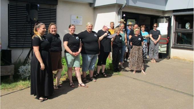 Foto einer Gruppe von Erwachsenen vor einem Gebäude an einem sonnigen Tag. Mehrere Personen stehen gemeinsam vor dem Eingang und wirken freundlich und zugewandt. Einige tragen schwarze Kleidung mit einem kleinen Logo, im Hintergrund sind Fenster, eine Tür und ein Schild am Gebäude zu sehen. Das Bild vermittelt Gemeinschaft, Begegnung und Engagement vor Ort.